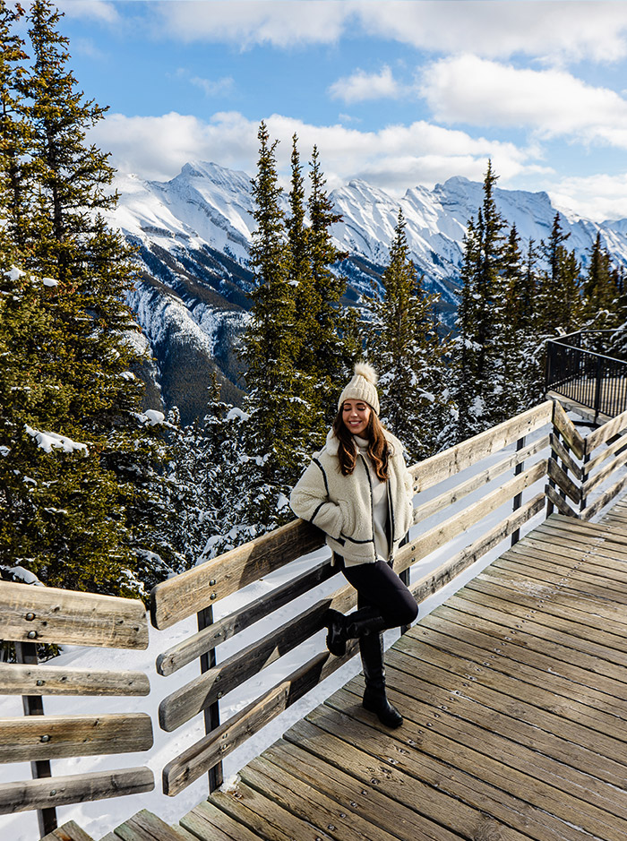 Kelsey standing on board walk on the top of a mountain with snow capped mountain ranges in the background