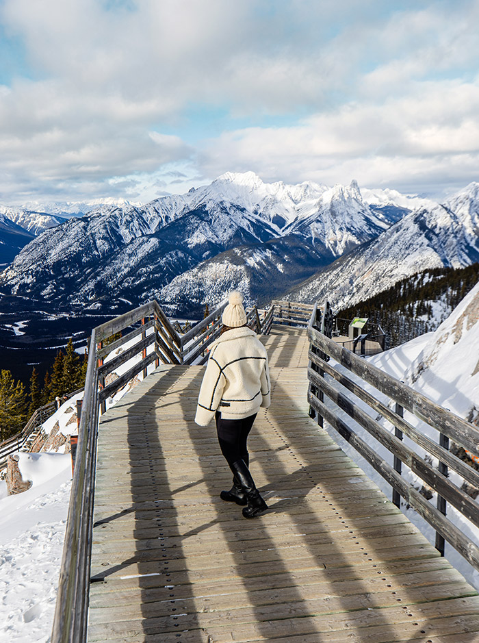 Kelsey walking along a boardwalk on top of a mountain range, snow capped mountains can be seen in background