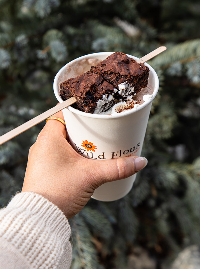 A hand holding a paper cup filled with hot chocolate, topped with a brownie and whipped cream