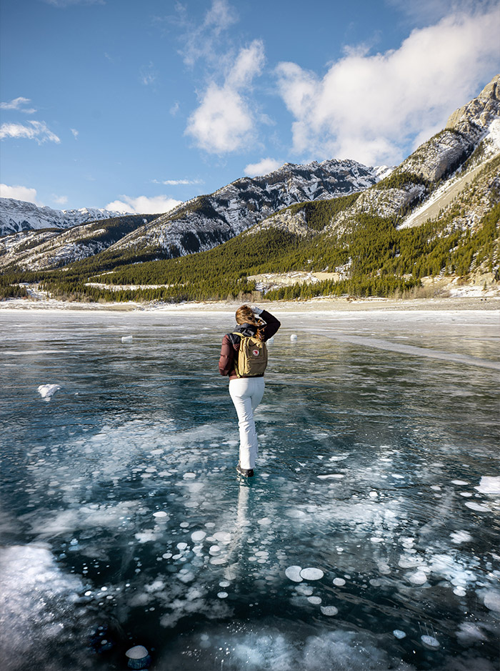 Kelsey walking across a frozen lake which is covered in ice bubbles, snow capped mountains can be seen in background