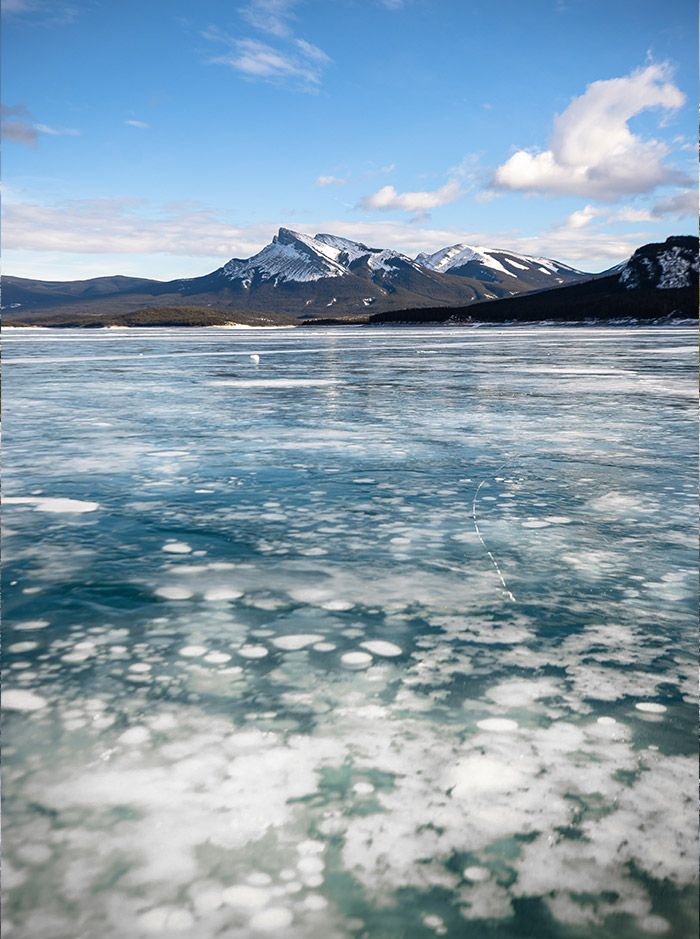 a frozen lake which is covered in ice bubbles, snow capped mountains can be seen in background