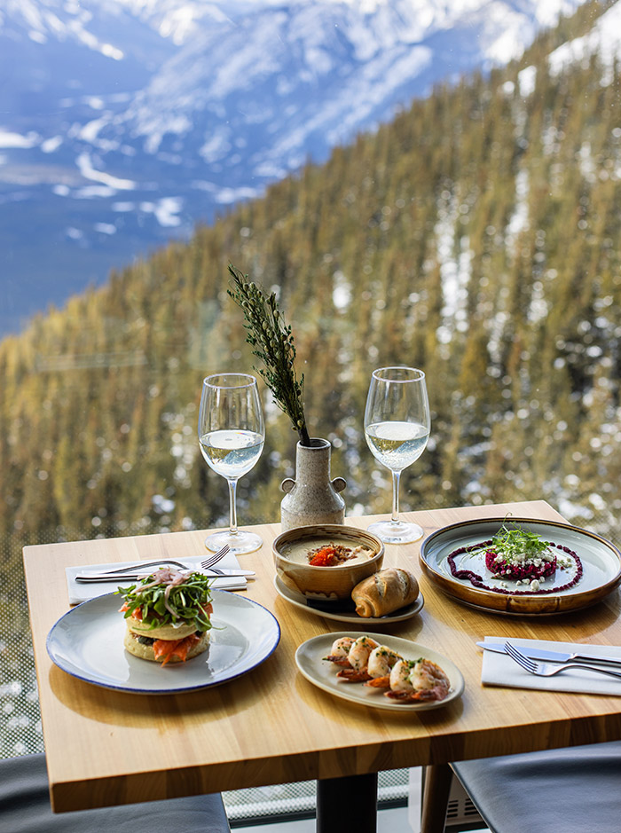A table with two glasses of wine and various dishes of food, behind the table is a glass window which shows a view of snow capped mountains 