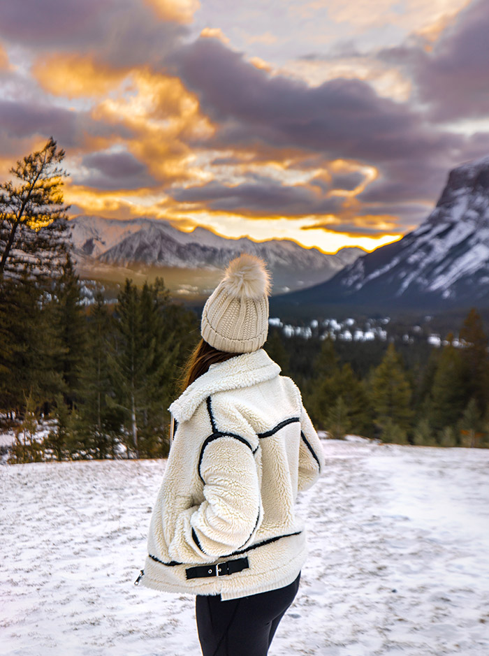 Kelsey wearing a beanie and winter jacket looking at a beautiful sunrise over snow capped mountains