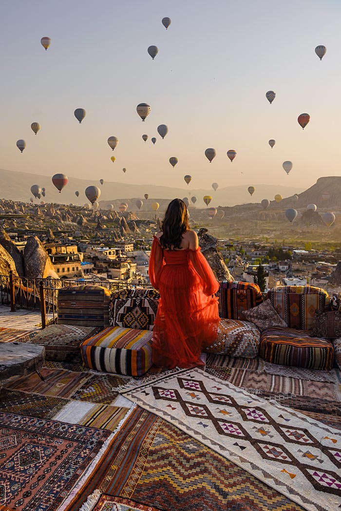 A woman in a red dress stands on a terrace adorned with colorful Turkish rugs, gazing at numerous hot air balloons floating over the rocky landscape of Cappadocia, Turkey, during sunrise