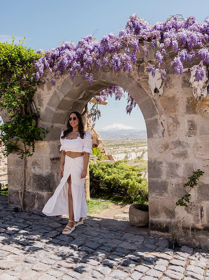A woman in a white two-piece outfit stands under a stone archway adorned with purple flowers, with a scenic view of the Cappadocia landscape and a snow-capped mountain in the background, at Argos Hotel in Cappadocia, Turkey.