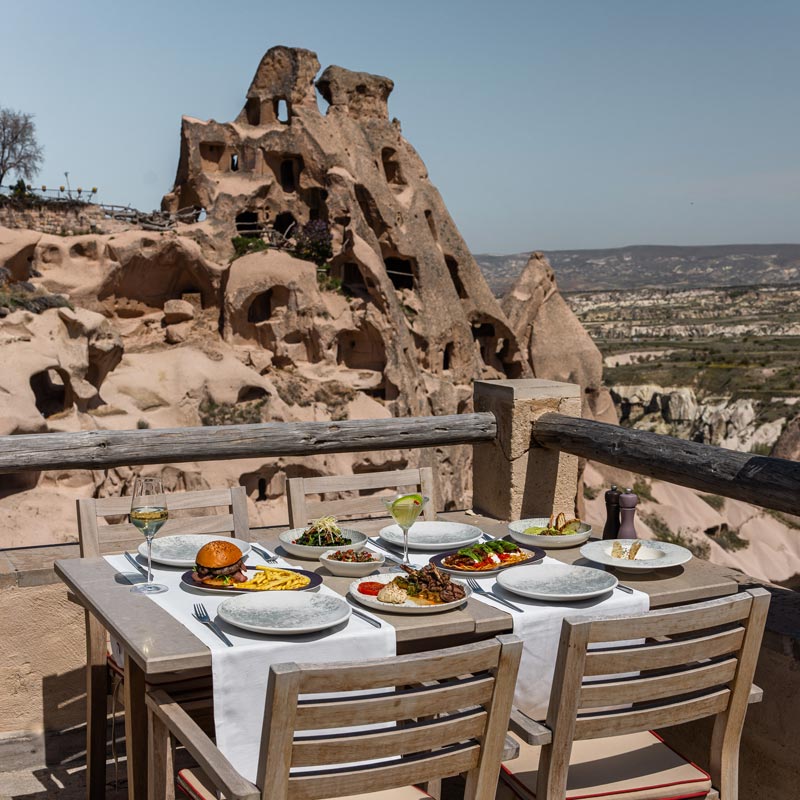 A scenic outdoor dining table set for a meal with multiple dishes and drinks, overlooking the unique rock formations of Cappadocia. The rustic stone structures and vast landscape create an enchanting backdrop for a memorable dining experience.

