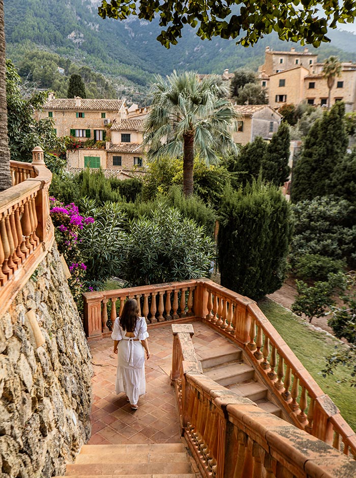A woman in a white dress descends a terracotta staircase at Belmond La Residencia in Mallorca, surrounded by lush greenery and vibrant bougainvillaea. The stone walls and balustrades add a rustic charm to the picturesque setting, with traditional stone houses and palm trees in the background. The serene landscape of the mountainous region enhances the tranquil and idyllic atmosphere.