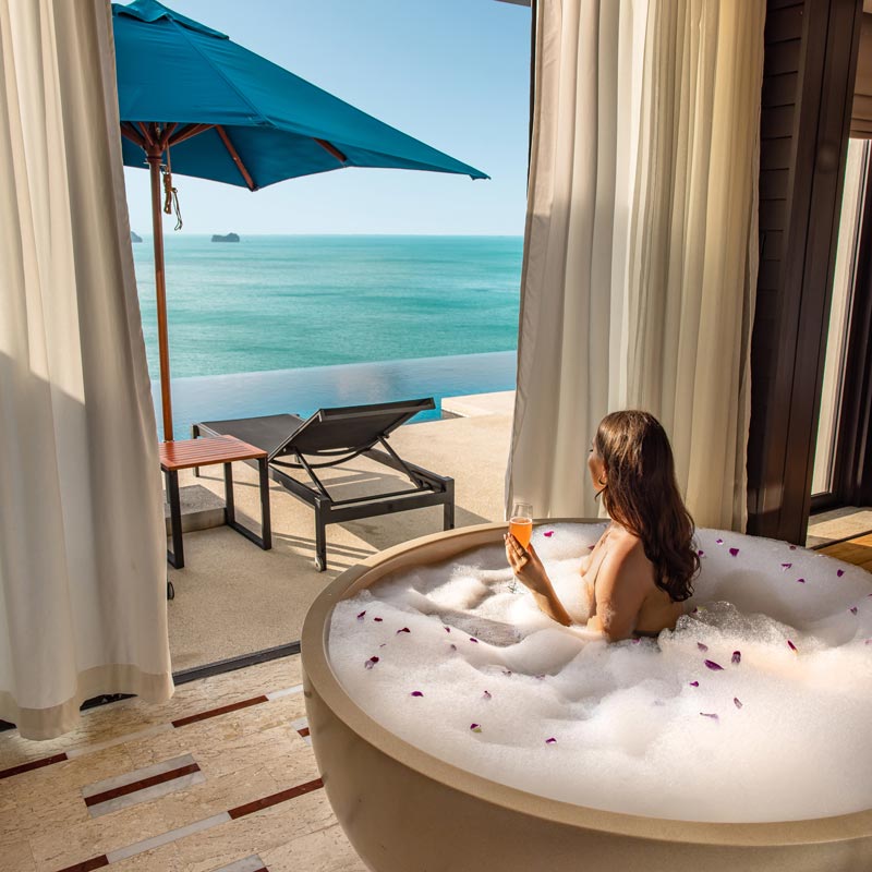 A woman relaxes in a bubble-filled outdoor bathtub, holding a drink while enjoying a breathtaking ocean view. The scene is framed by white curtains and features a sun lounger and a blue umbrella on a terrace, with the infinity pool blending seamlessly into the horizon. The tranquil setting epitomizes luxury and serene coastal living.






