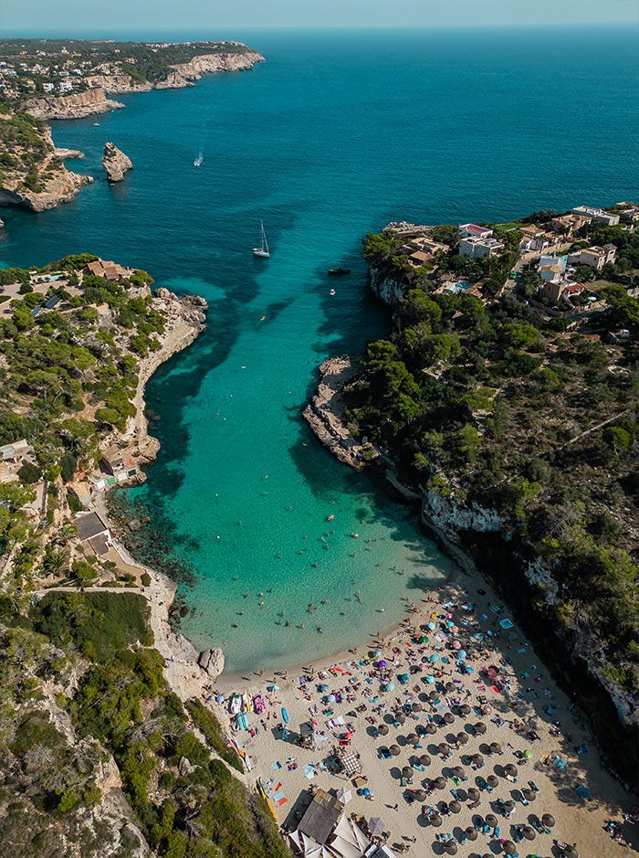 A breathtaking aerial view of Cala Llombards in Mallorca, showcasing a stunning cove with turquoise waters surrounded by lush greenery and rocky cliffs. The sandy beach below is dotted with colorful umbrellas and sunbathers enjoying the serene environment. In the cove, several people swim and boats float, highlighting the picturesque and inviting nature of this beautiful location.
