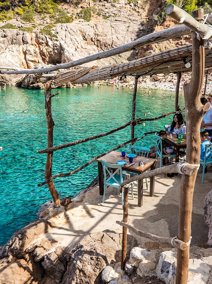 A rustic, seaside dining area at Ca's Patró March in Mallorca features simple wooden tables and blue chairs set against a stunning backdrop of clear, turquoise waters. The natural setting is enhanced by rough-hewn wooden railings and a canopy of woven reeds, providing a shaded spot to enjoy a meal while surrounded by rocky cliffs and the serene beauty of the cove. People are seen dining and relaxing, adding to the laid-back atmosphere of this picturesque location.