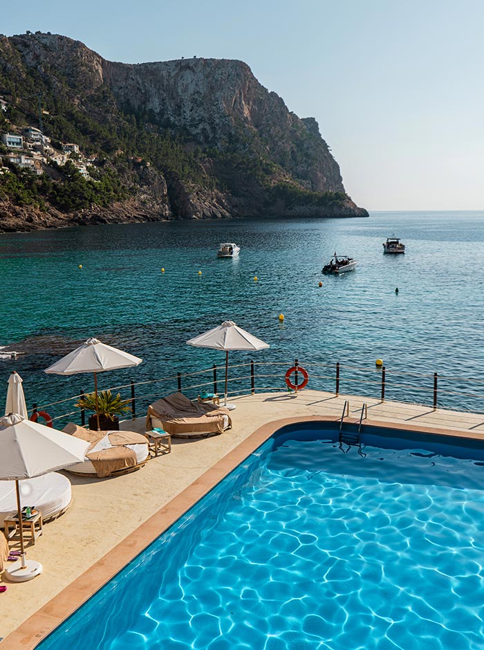 View from Gran Folies Beach Club in Mallorca, showcasing a stunning infinity pool with clear blue water and luxurious sun loungers under white umbrellas. The pool area overlooks a serene bay with anchored boats and a backdrop of dramatic cliffs covered in greenery, offering a breathtaking coastal experience.