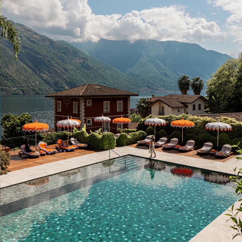 A picturesque pool area at a luxury hotel features vibrant orange and white umbrellas and cushioned lounge chairs, set against the backdrop of a serene lake and towering green mountains. The scene includes charming red and white buildings and lush greenery, with a woman in a summer dress and hat walking by the pool. The idyllic setting exudes relaxation and natural beauty.






