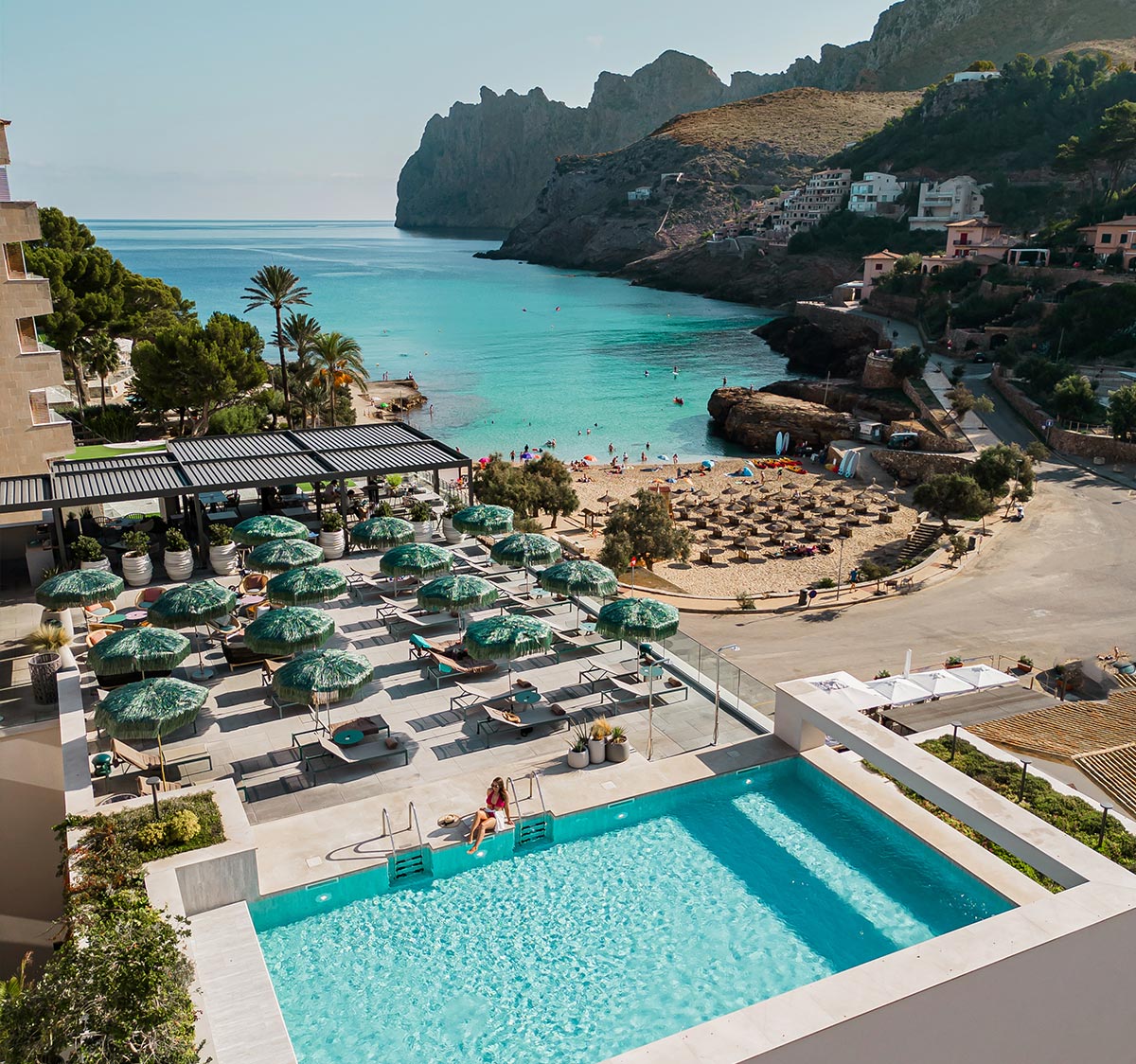 This image depicts a luxury hotel with a stunning view of a coastal area featuring turquoise waters, a sandy beach with sun loungers, and surrounding cliffs. The foreground showcases a rooftop pool and lounge area with shaded sunbeds, where a guest is seated at the edge of the pool. The idyllic setting combines modern elegance with natural beauty.