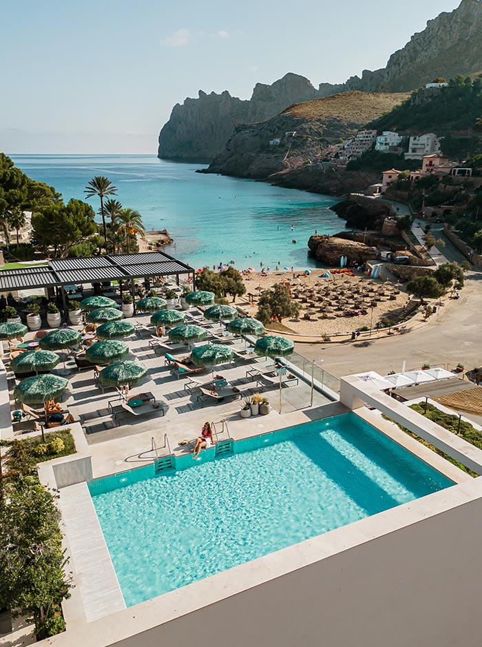 A scenic view of Hotel El Vicenç de la Mar in Mallorca, showcasing a pristine swimming pool with a woman relaxing by the edge, overlooking the clear blue waters of a beautiful bay. The hotel features an array of shaded loungers with green umbrellas, a sandy beach with thatched parasols, and dramatic cliffs in the background, creating a perfect blend of luxury and natural beauty.






