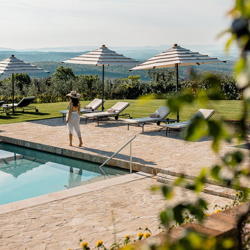 A serene poolside scene at a luxury resort in Tuscany, featuring sun loungers and large umbrellas set on a stone terrace. A woman in a white dress and sun hat walks beside the clear blue pool, with a backdrop of rolling hills and lush greenery. The peaceful landscape offers expansive views of the countryside, providing a relaxing and picturesque setting.






