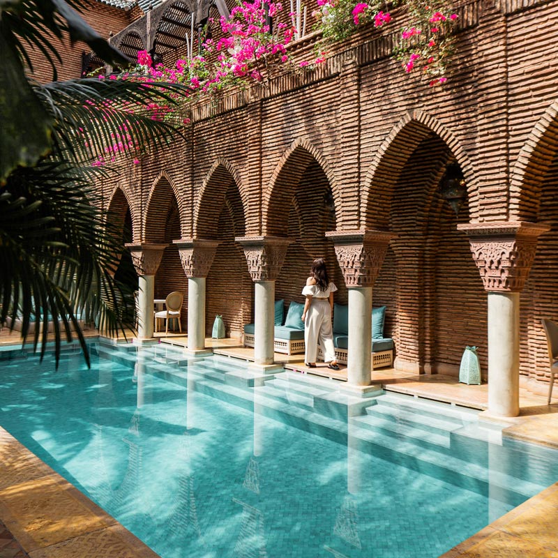 A luxurious courtyard featuring a turquoise pool lined with stone columns supporting brick arches. Pink bougainvillea flowers cascade from the upper level, adding a splash of color. A woman in a white dress stands by the pool, with plush seating and turquoise cushions beneath the arches, creating a serene and elegant atmosphere.
