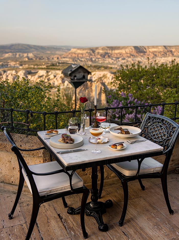 A romantic dining table set for two with gourmet dishes, wine glasses, and a single red rose in a vase, overlooking the picturesque landscape of Cappadocia, Turkey, at Lil'a Restaurant in the Museum Hotel.