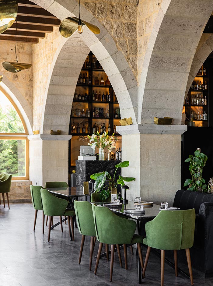 An elegant restaurant interior with arched stone ceilings, green upholstered chairs, and a well-stocked bar, featuring modern lighting and large windows allowing natural light, at Millocal in Cappadocia, Turkey.