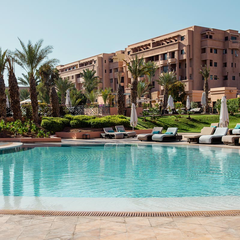 A luxurious outdoor pool area at a resort, featuring crystal-clear water surrounded by comfortable lounge chairs and umbrellas. The background showcases a large, modern building with terracotta hues and lush landscaping, including palm trees and well-manicured shrubs. The scene is set against a clear blue sky, creating a perfect setting for relaxation and leisure.







