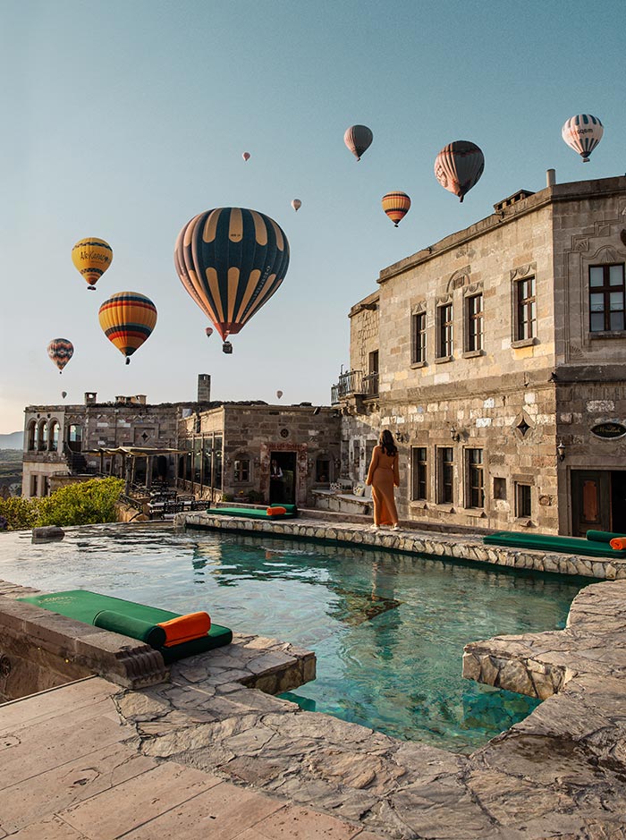 A woman in an orange dress walks by a stone pool with lounge chairs, overlooking numerous hot air balloons floating in the sky above the historic stone buildings of the Museum Hotel in Cappadocia, Turkey.