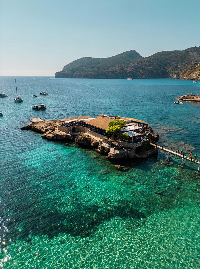 Aerial view of Restaurante Illeta in Mallorca, a charming restaurant situated on a rocky outcrop surrounded by crystal-clear turquoise waters. The eatery features an open-air dining area with a thatched roof, connected to the mainland by a narrow walkway. The backdrop includes picturesque mountains and several anchored boats, highlighting the serene and idyllic setting of this coastal dining spot.