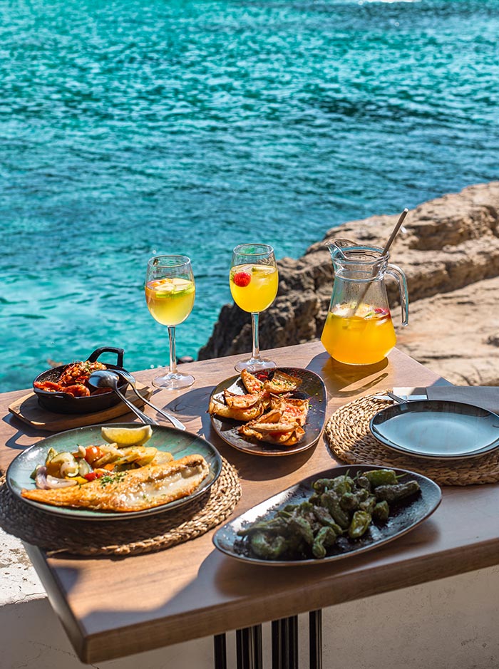 A delightful meal setup at Restaurante Illeta in Mallorca, featuring a variety of Mediterranean dishes on a wooden table by the sea. The spread includes a grilled fish fillet with lemon wedges and vegetables, a plate of roasted potatoes, a bowl of padron peppers, and a dish of shrimp in a red sauce. Two glasses of refreshing sangria with fruit slices and a pitcher of sangria sit next to the food. The stunning turquoise waters and rocky coastline create a beautiful and relaxing dining backdrop.