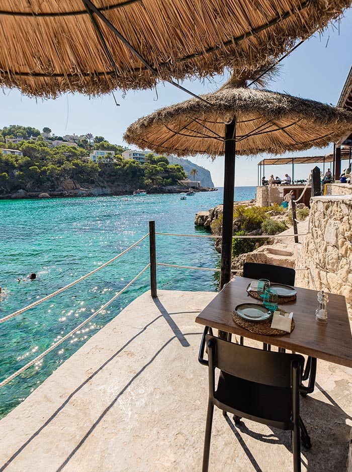 A serene seaside dining experience at Restaurante Illeta in Mallorca, featuring a thatched umbrella providing shade over a table set for two with rustic wooden chairs. The table overlooks crystal-clear turquoise waters and the picturesque coastline dotted with lush greenery and cliffs. Swimmers can be seen enjoying the water, while the distant view includes charming seaside homes and boats gently floating in the cove, creating a perfect backdrop for a relaxing meal.