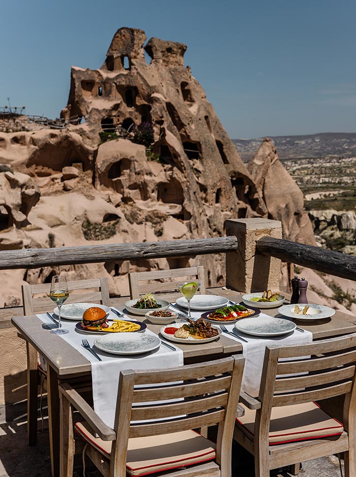 An outdoor dining table set with various dishes and drinks, overlooking the unique rock formations and cave dwellings of Cappadocia, Turkey, at Seki Restaurant in the Argos Hotel.