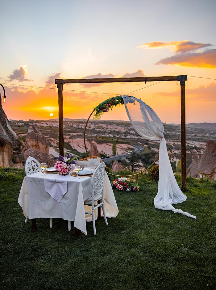 A romantic outdoor dining setup with a table for two, adorned with flowers and wine glasses, under a decorated archway overlooking the rocky landscape of Uçhisar, Cappadocia, at sunset.