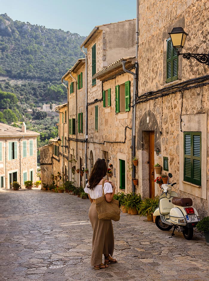 A charming cobblestone street in Valldemossa, Mallorca, lined with traditional stone houses featuring green shutters and potted plants. A woman in a white top and beige skirt, carrying a woven bag, stands admiring the picturesque scene with lush green mountains in the background. A classic white scooter is parked to the right, enhancing the quaint village atmosphere.