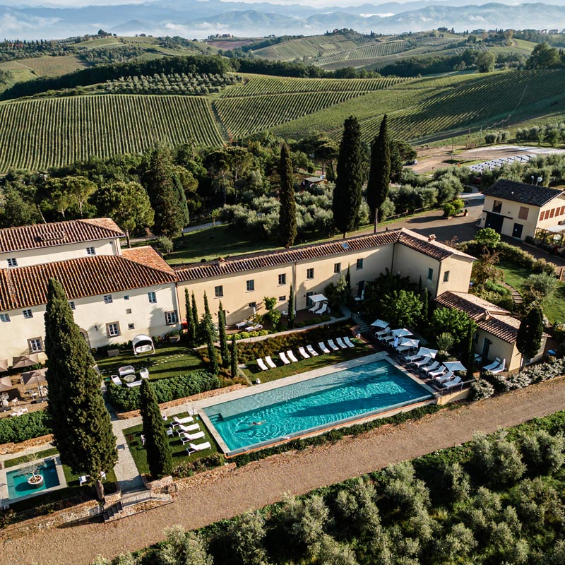 An aerial view of a luxurious villa in Tuscany surrounded by lush vineyards and rolling hills. The villa features a long, rectangular pool with sun loungers and umbrellas, set amidst well-manicured gardens and tall cypress trees. The picturesque landscape includes terracotta-roofed buildings and expansive views of the serene countryside, evoking a sense of tranquility and elegance.







