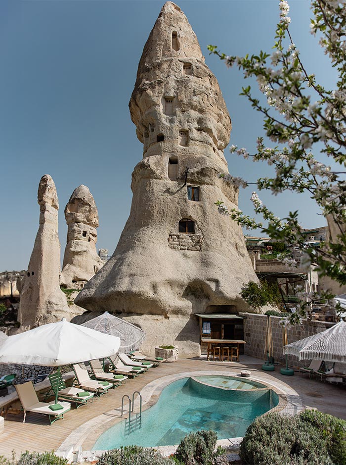 A unique rock formation with windows carved into it stands next to an outdoor pool area with lounge chairs, umbrellas, and a small bar at Aza Cave Hotel in Cappadocia, Turkey. The sky is clear, and blossoming trees add to the scenic view.