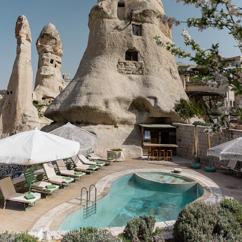 A charming pool area nestled among the distinctive fairy chimneys of Cappadocia. The setting includes sun loungers, umbrellas, and hammocks, offering a relaxing retreat with the historic rock formations towering above.

