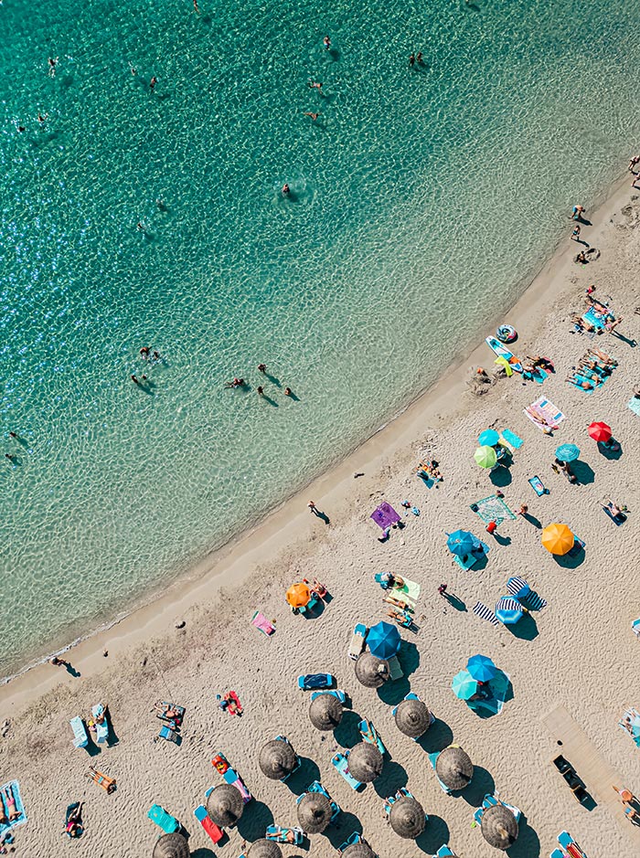 Aerial view of a vibrant beach in Mallorca, showing clear turquoise waters and a sandy shore filled with colorful beach umbrellas and sunbathers. The shallow water allows for easy swimming, while rows of straw parasols provide shade for those relaxing on the beach. The scene captures the lively and inviting atmosphere of one of Mallorca's best beaches.


