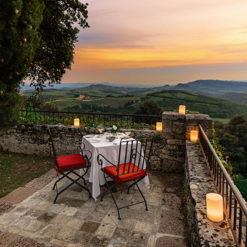 A picturesque outdoor dining setup on a stone patio overlooking a rolling Tuscan landscape at sunset. The table is elegantly set with white linens and red-cushioned chairs, surrounded by soft candlelight. The scene captures a romantic and serene atmosphere with expansive views of green hills and a colorful sky.






