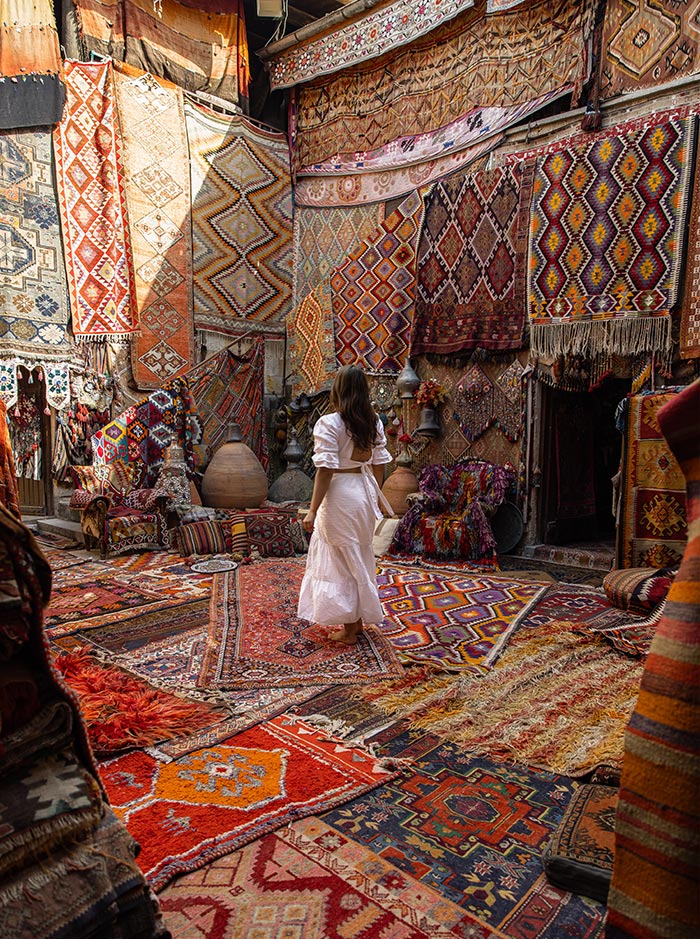 A woman in a white dress walking through a vibrant marketplace filled with a variety of colorful, patterned Turkish rugs hanging on the walls and spread on the floor at Galerie Ikman in Cappadocia, Turkey