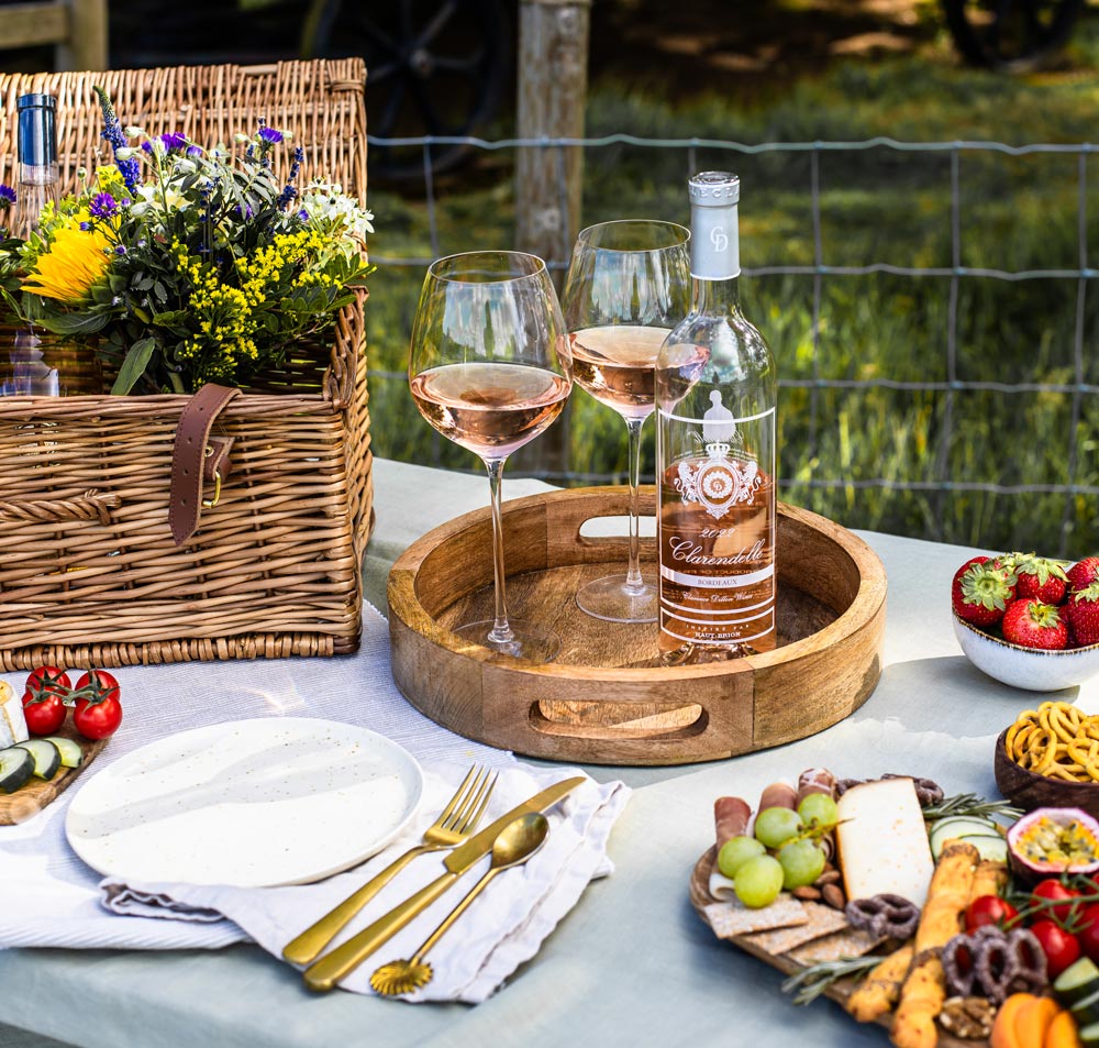 A beautifully arranged picnic setup featuring a wicker basket filled with flowers, a bottle of rosé wine with two filled glasses on a wooden tray, and a variety of fresh fruits, cheese, and snacks on a table. The background shows a grassy area with a fence, suggesting an outdoor setting