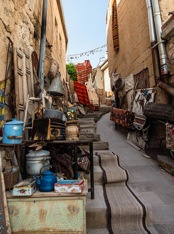 A narrow alleyway lined with various antiques and traditional Turkish rugs for sale, leading up a small incline between stone buildings in Göreme, Cappadocia, Turkey.