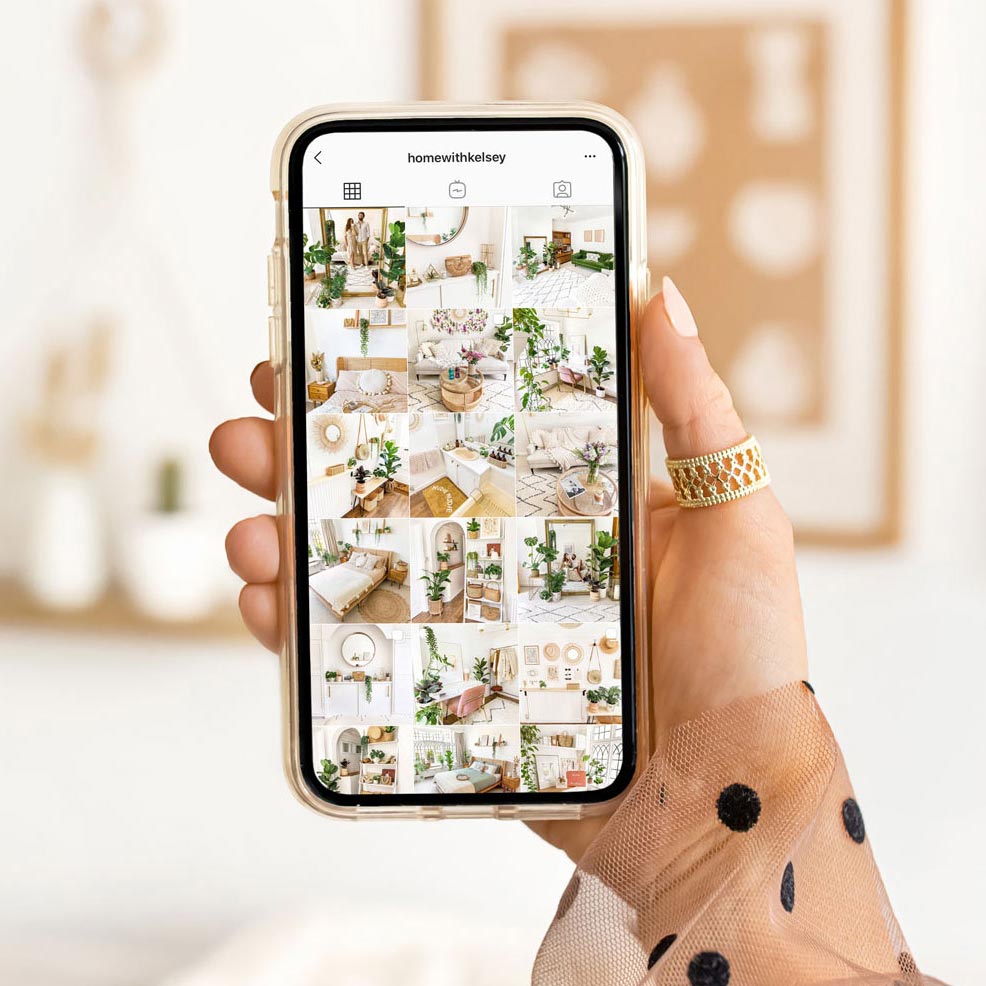 A hand holding a smartphone displaying an Instagram profile named 'homewithkelsey,' showcasing a grid of aesthetically pleasing home decor images. The background is a blurred interior setting, highlighting the focus on the phone screen