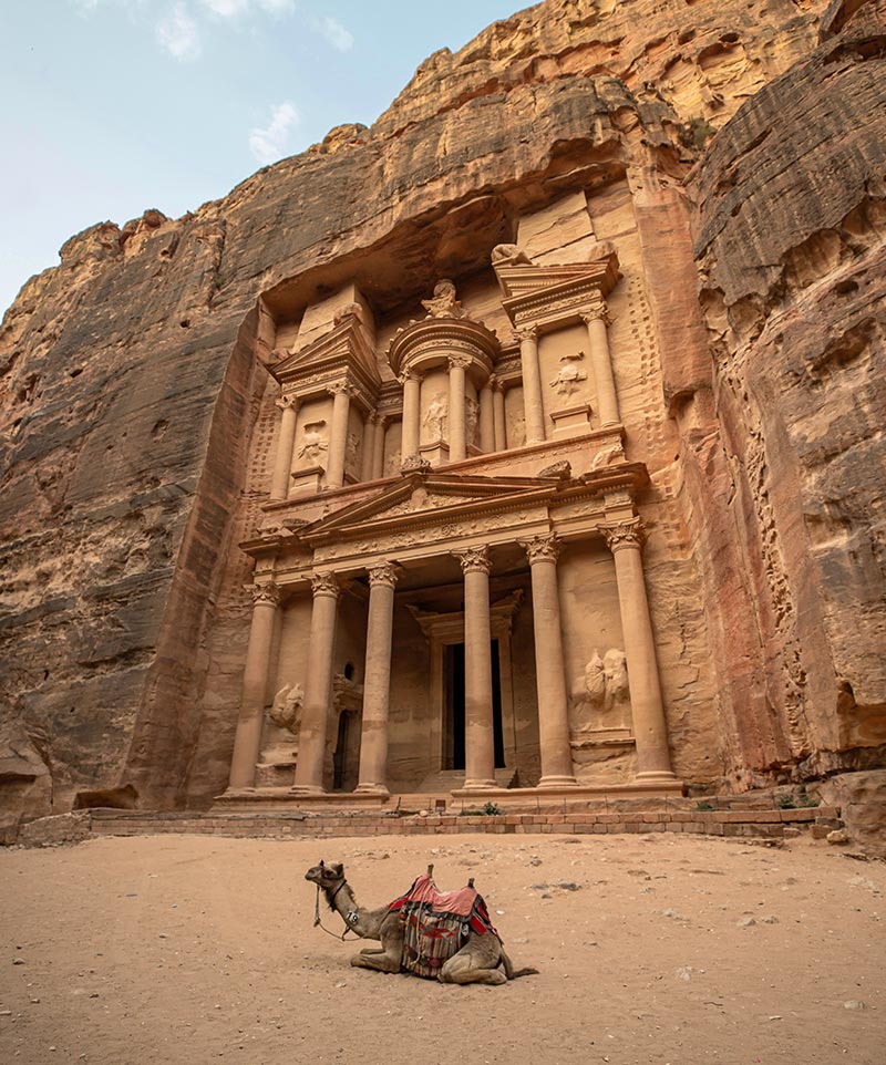 The iconic rock-carved facade of Al-Khazneh (The Treasury) in Petra, Jordan, with a camel resting in the foreground under a clear sky
