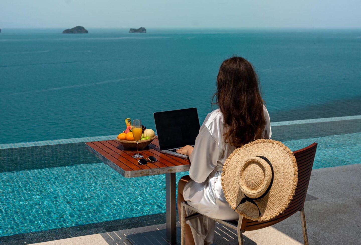 A woman wearing a white robe sits at a table by an infinity pool overlooking the ocean, working on a laptop. A straw hat rests on the back of her chair, and a bowl of fresh fruit and a glass of juice are on the table, suggesting a relaxing yet productive setting
