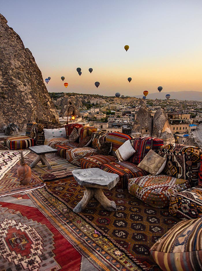 A terrace adorned with colorful Turkish rugs and cushions, offering a cozy seating area with wooden tables, overlooking hot air balloons floating over the rocky landscape of Cappadocia, Turkey, at sunrise