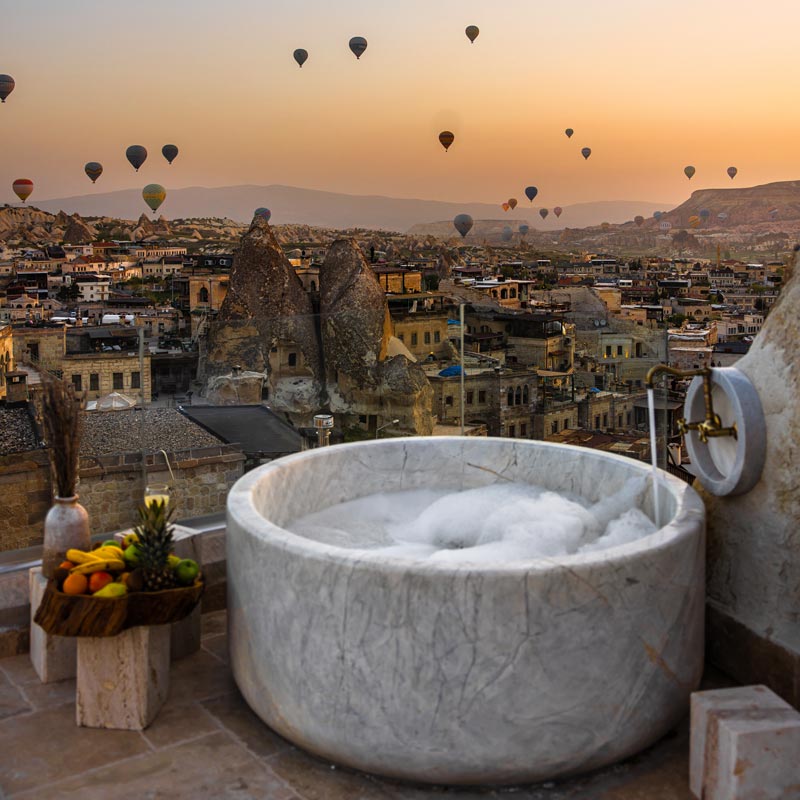 A luxurious outdoor marble bathtub filled with bubbles offers a stunning view of Cappadocia's unique rock formations and fairy chimneys at sunset. The sky is dotted with numerous hot air balloons floating above the historic town. A tray of fresh fruit is placed nearby, enhancing the serene and picturesque atmosphere.


