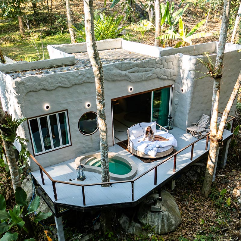 A modern villa nestled in the woods, featuring a unique stone facade and large windows. A woman lounges on a circular daybed on the wooden deck, which includes a small plunge pool. The villa is surrounded by lush greenery, creating a tranquil and immersive natural retreat.







