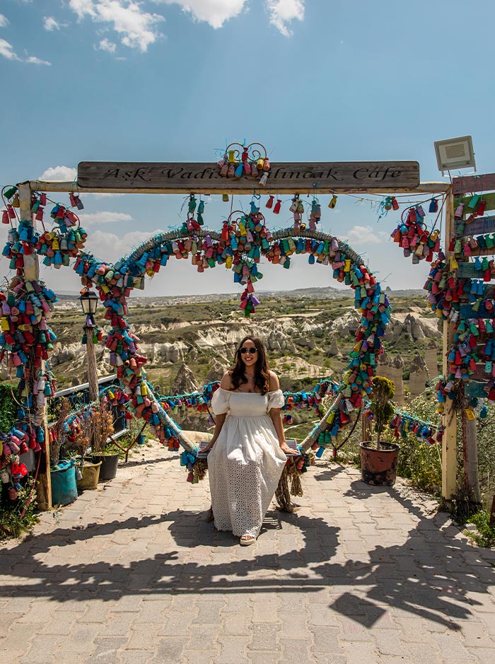 A woman in a white dress sits on a swing adorned with colorful decorations, positioned under a sign that reads 'Aşk Vadisi Salıncak Cafe,' with the scenic landscape of Love Valley, Cappadocia, Turkey, in the background