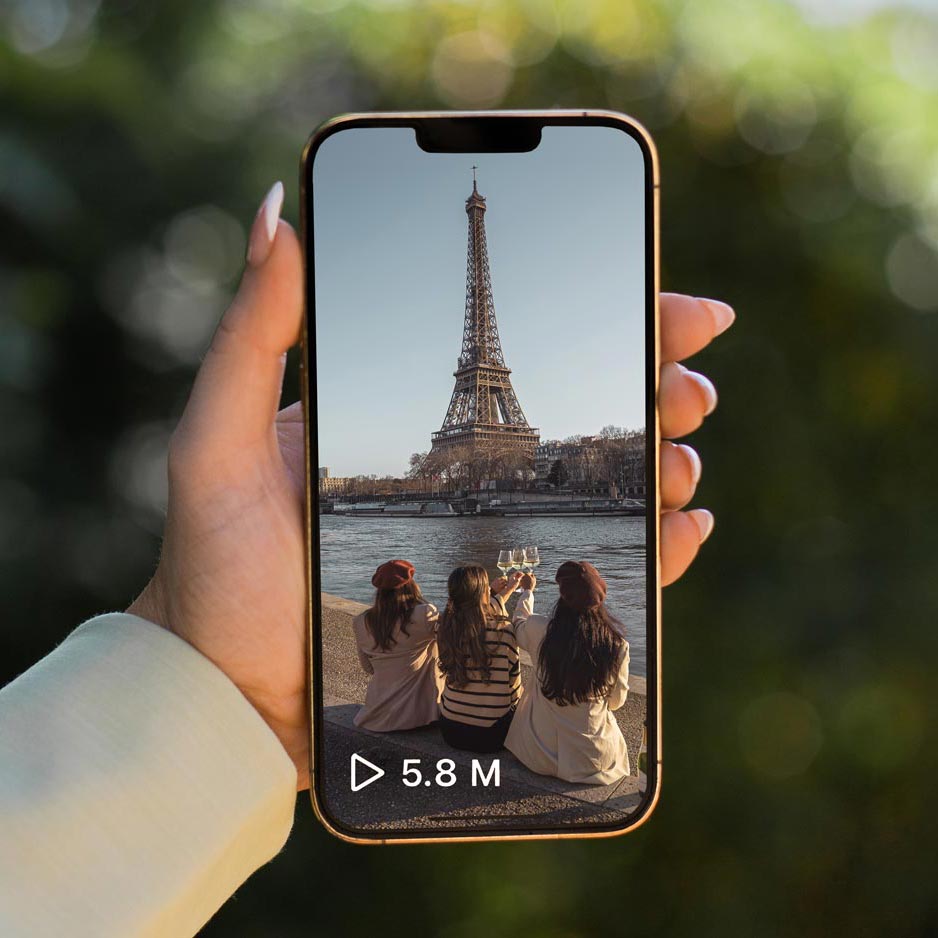 A hand holding a smartphone displaying a popular video with 5.8 million views, showing three women sitting by the Seine River toasting with glasses of wine, with the Eiffel Tower in the background in Paris, France.