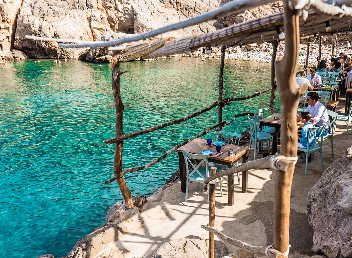 This image shows a picturesque seaside dining area at a rustic restaurant with wooden tables and light blue chairs, overlooking clear turquoise water. The space is shaded by a natural wooden structure, while diners enjoy the scenic view and the relaxed coastal atmosphere. Swimmers can be seen in the background, adding to the idyllic setting.