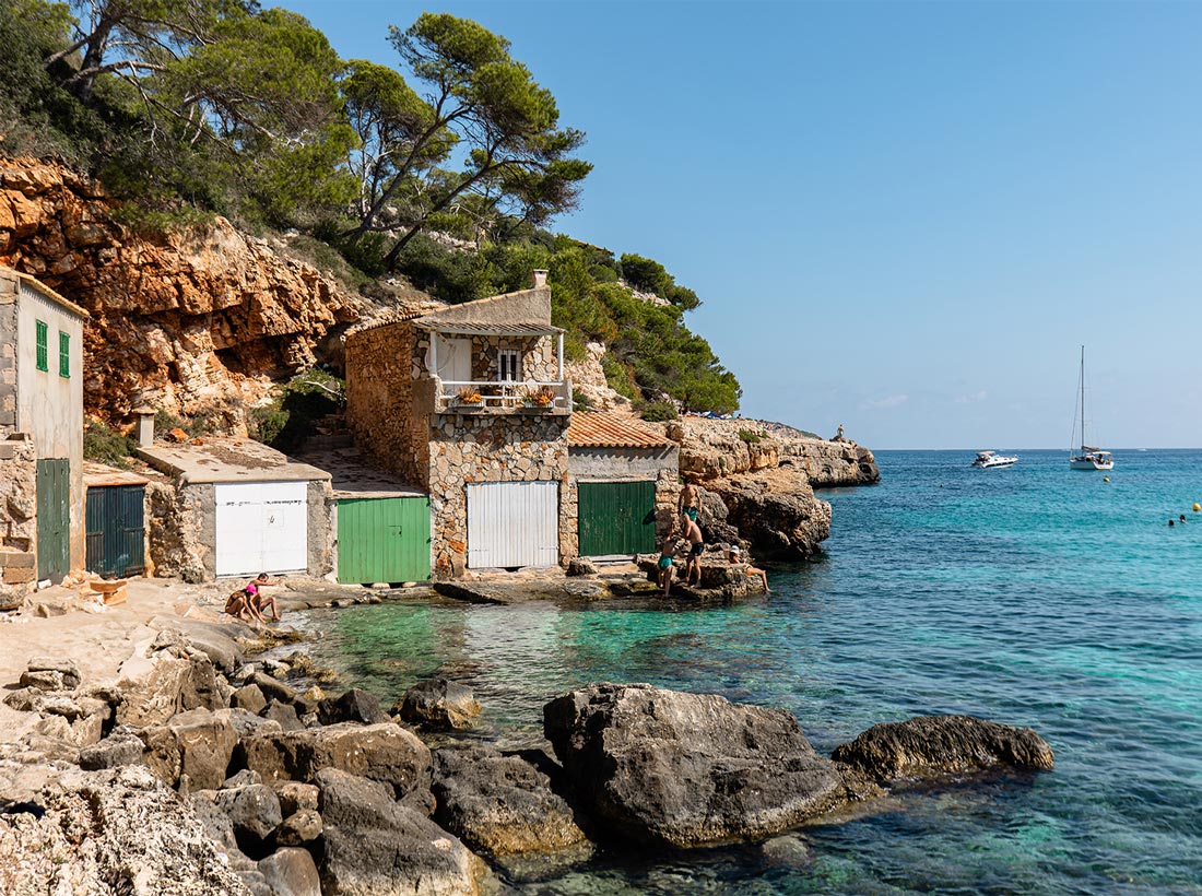 A picturesque coastal scene in Cala Llombards, Mallorca, featuring rustic stone houses with green and white doors built into the rocky shoreline, crystal-clear turquoise waters, and boats anchored in the distance under a bright blue sky.