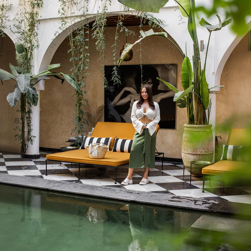 A cozy courtyard with a checkered tile floor features a pool and vibrant greenery hanging from arches. A woman in a white top and green skirt stands beside a mustard-yellow lounge chair with black and white striped pillows. The setting is lush and inviting, accented by a large potted plant and a woven basket on the chair.







