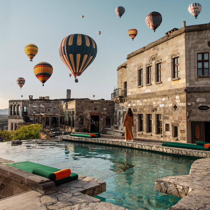 A luxurious pool area at sunset, surrounded by historic stone buildings and numerous hot air balloons floating in the sky. A woman in a long dress stands by the pool, which reflects the vibrant colors of the balloons and the evening light, creating a magical and picturesque scene.






